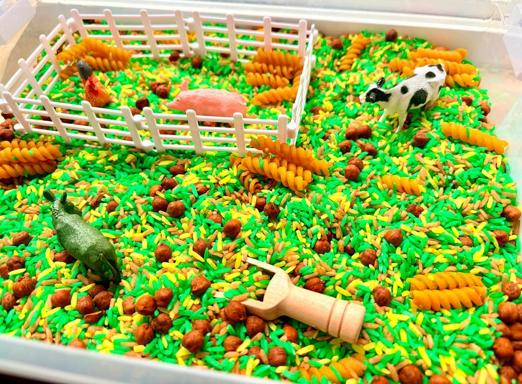 A sensory play tray filled with bright green and yellow rice, brown pellets, orange corn-shaped pieces, a white fence, a small pink pig, a cow figurine, a donkey figurine, and a wooden scoop.