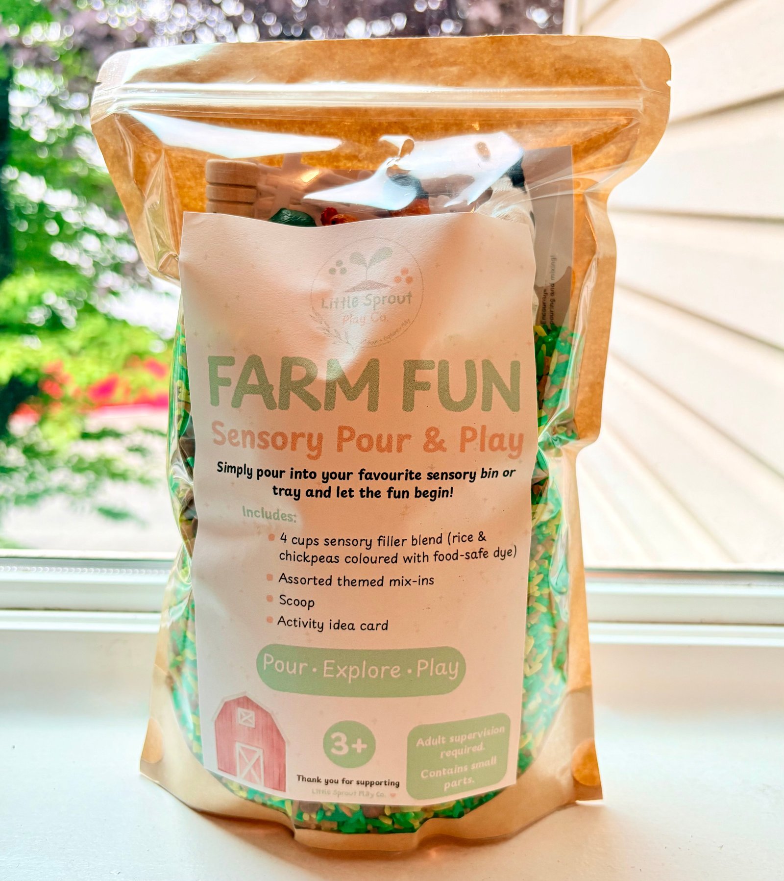 A bag of LittleSproutPlayCo Farm Fun Sensory Pour & Play Sensory Kit, featuring a white label with green and orange text, set on a windowsill with a blurred garden background.