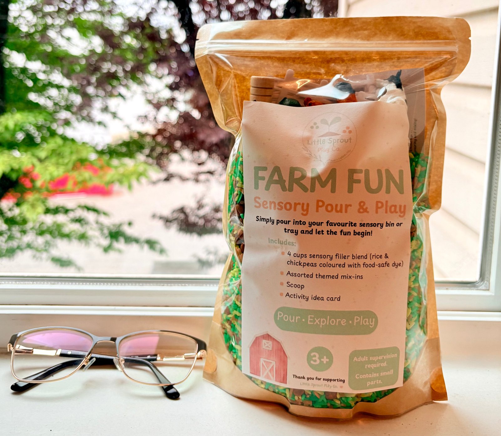 A stand-up pouch of 'Farm Fun' Sensory Pour & Play Sensory Play Kit by LittleSproutPlayCo. sits on a windowsill with a pair of glasses to show size comparison