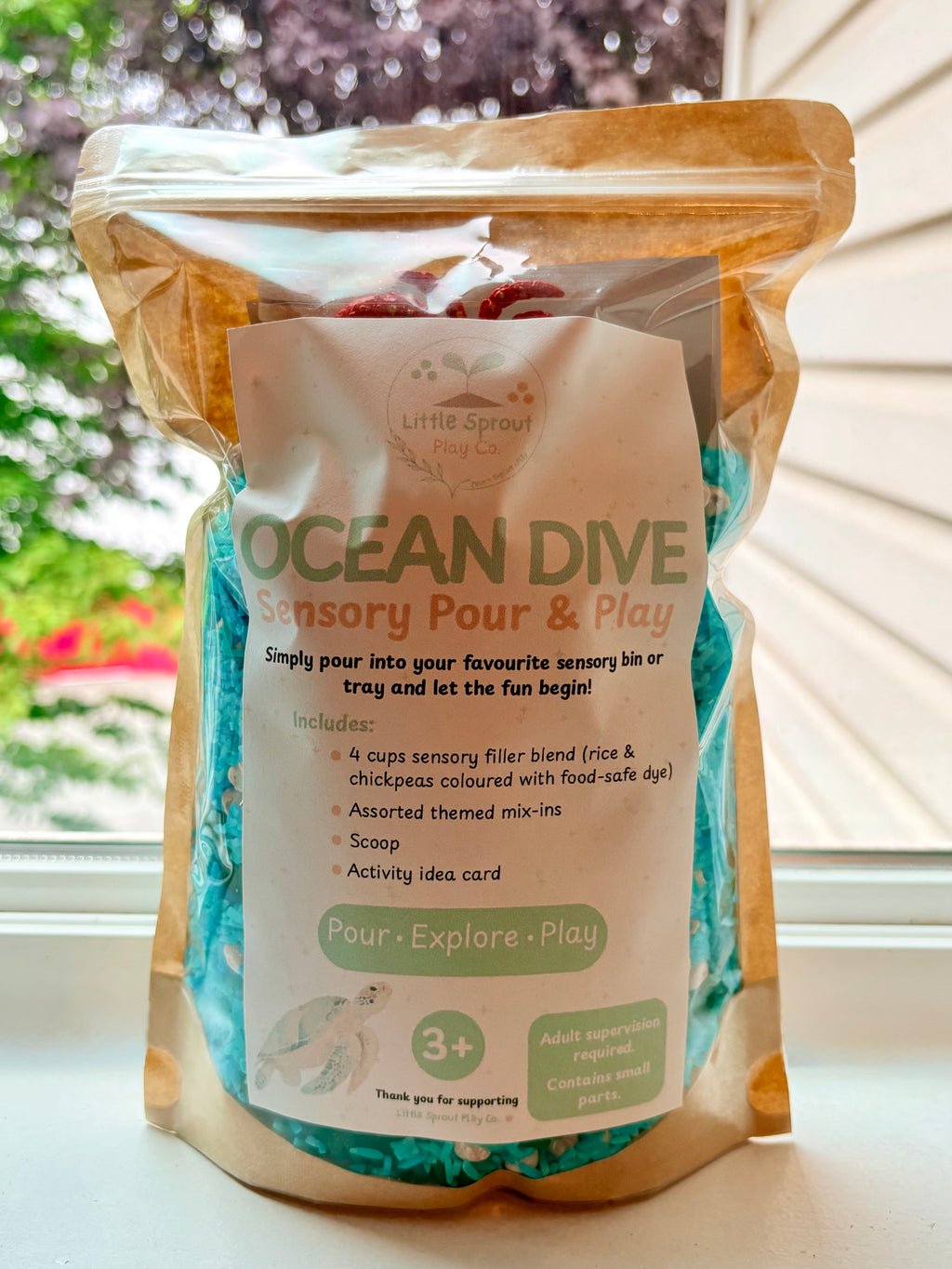 A stand-up resealable pouch of  LittleSproutPlayCo's  'Ocean Dive' Sensory Pour & Play Sensory Kit, filled with blue and green textured filler, set on a windowsill with a blurred outdoor background.