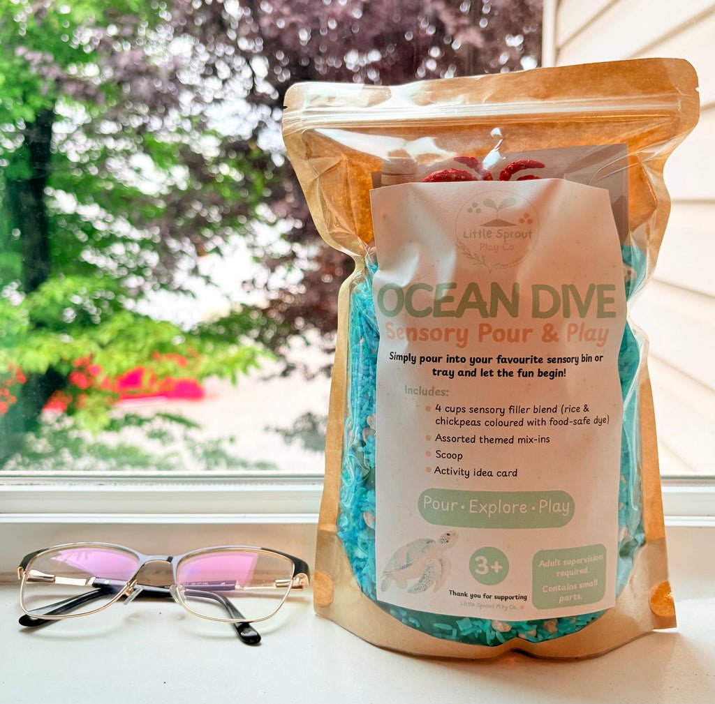 A stand-up pouch of 'LittleSproutPlayCo's Ocean Dive Sensory Pour & Play sensory filler sitting beside glasses. The background shows a blurred green garden view through the window.