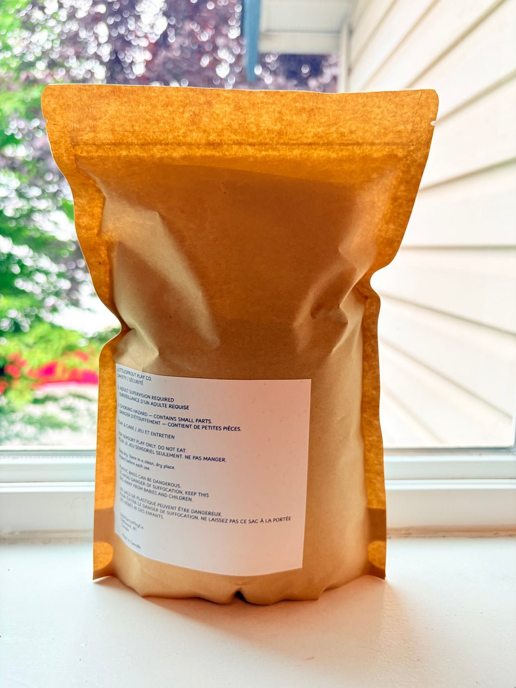 A stand-up kraft paper pouch with a white label, photographed on a windowsill with a window and outdoor greenery in the background.